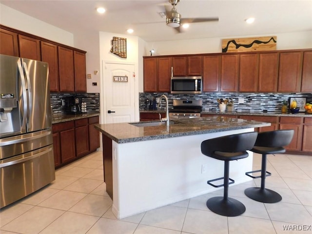 kitchen with stainless steel appliances, a kitchen island with sink, a sink, light tile patterned flooring, and dark stone countertops