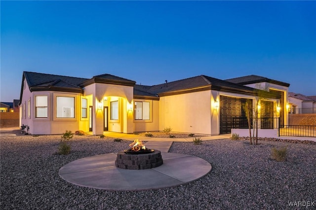 back of property at dusk featuring an attached garage, fence, a fire pit, and stucco siding