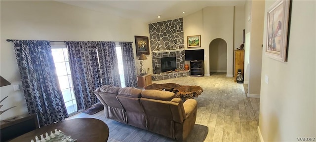 living area featuring baseboards, arched walkways, wood finished floors, a stone fireplace, and high vaulted ceiling