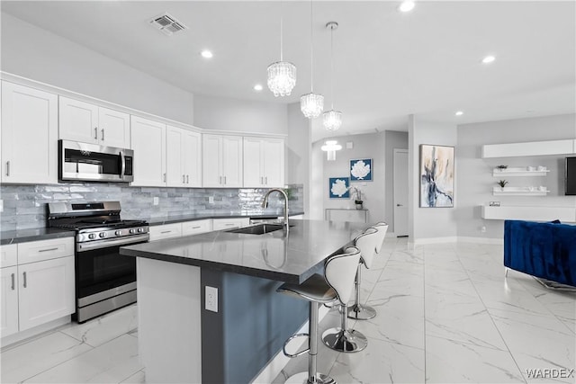 kitchen with a center island with sink, dark countertops, hanging light fixtures, stainless steel appliances, and a sink