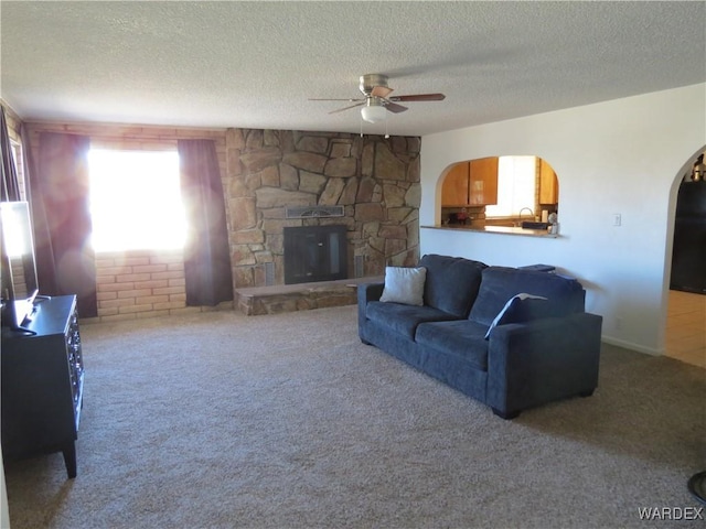 living area featuring arched walkways, ceiling fan, carpet, a textured ceiling, and a stone fireplace