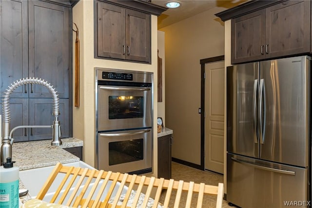 kitchen with light stone countertops, baseboards, appliances with stainless steel finishes, and dark brown cabinets