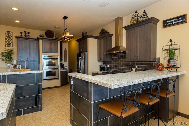 kitchen featuring light tile patterned floors, hanging light fixtures, stainless steel appliances, wall chimney range hood, and backsplash