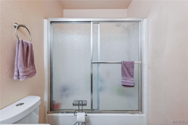 full bathroom featuring toilet, shower / bath combination with glass door, and a textured wall