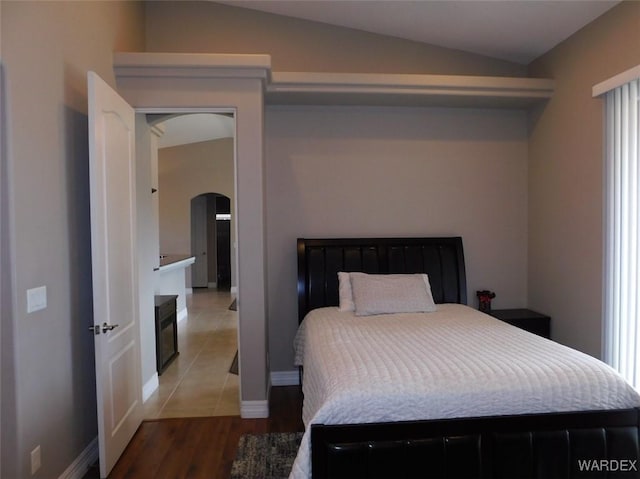 bedroom with baseboards, light wood-type flooring, lofted ceiling, a fireplace, and arched walkways