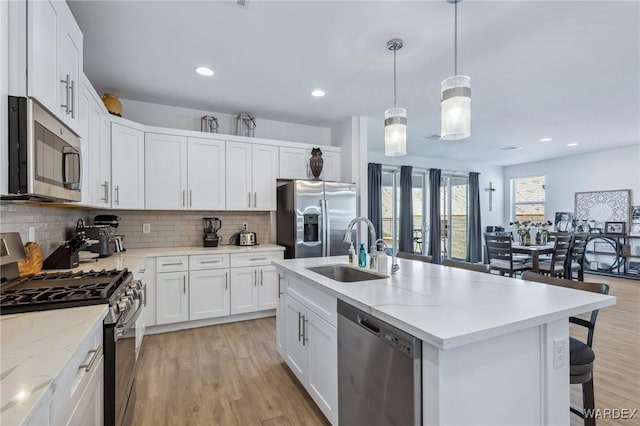 kitchen with a kitchen island with sink, stainless steel appliances, a sink, white cabinetry, and pendant lighting