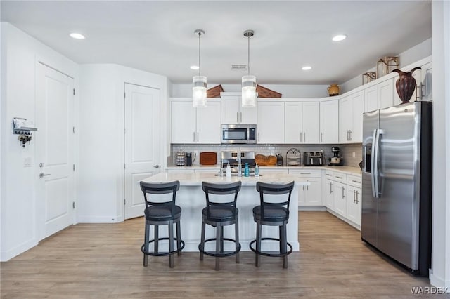 kitchen with light wood finished floors, a center island with sink, white cabinets, appliances with stainless steel finishes, and pendant lighting