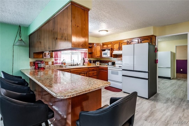 kitchen with white appliances, a peninsula, a sink, under cabinet range hood, and brown cabinets