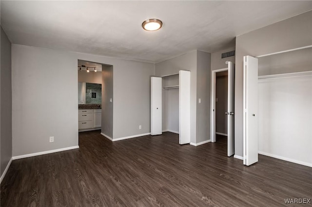 unfurnished bedroom featuring visible vents, baseboards, ensuite bath, dark wood-style flooring, and two closets