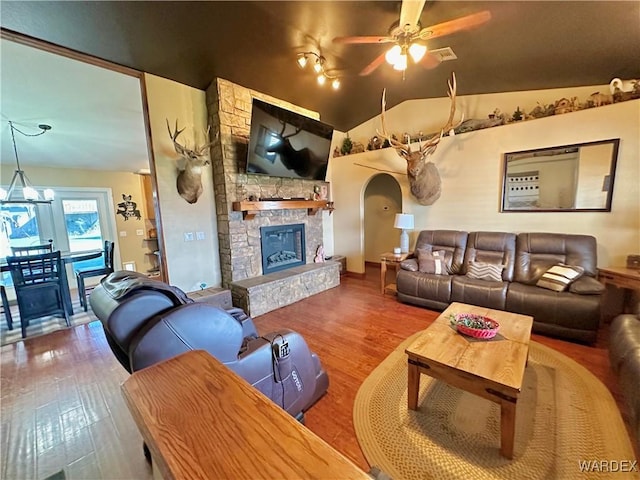 living area featuring arched walkways, vaulted ceiling, a stone fireplace, wood finished floors, and ceiling fan with notable chandelier