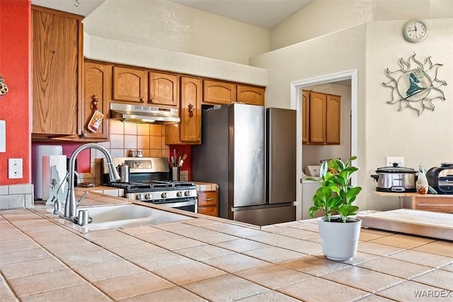 kitchen featuring tile countertops, brown cabinetry, appliances with stainless steel finishes, under cabinet range hood, and backsplash