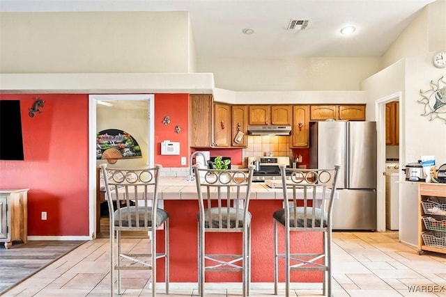 kitchen with visible vents, under cabinet range hood, tile countertops, a breakfast bar area, and appliances with stainless steel finishes