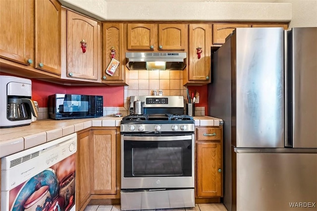 kitchen featuring under cabinet range hood, tile countertops, appliances with stainless steel finishes, and backsplash