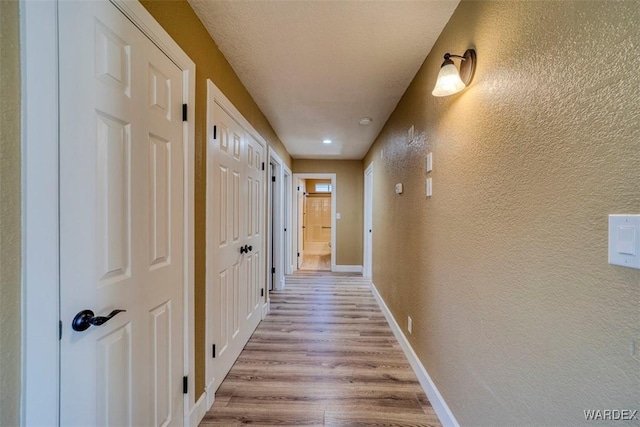 hallway featuring a textured ceiling, a textured wall, light wood-style flooring, and baseboards