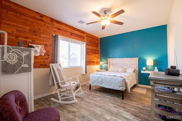 bedroom with ceiling fan, wood walls, wood finished floors, visible vents, and baseboards