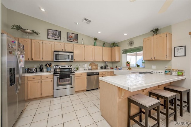 kitchen featuring a breakfast bar area, stainless steel appliances, visible vents, light brown cabinetry, and a peninsula