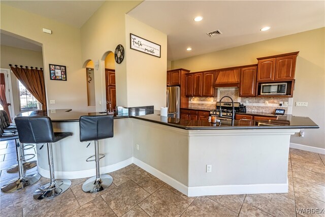 kitchen with a breakfast bar, dark countertops, custom range hood, appliances with stainless steel finishes, and a peninsula