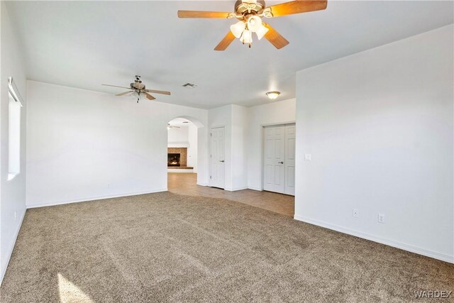 carpeted empty room with arched walkways, visible vents, a ceiling fan, a warm lit fireplace, and baseboards