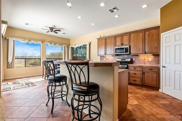 kitchen featuring visible vents, decorative backsplash, appliances with stainless steel finishes, a kitchen breakfast bar, and brown cabinetry