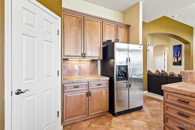 kitchen featuring brown cabinetry, stainless steel fridge with ice dispenser, light tile patterned floors, decorative backsplash, and arched walkways