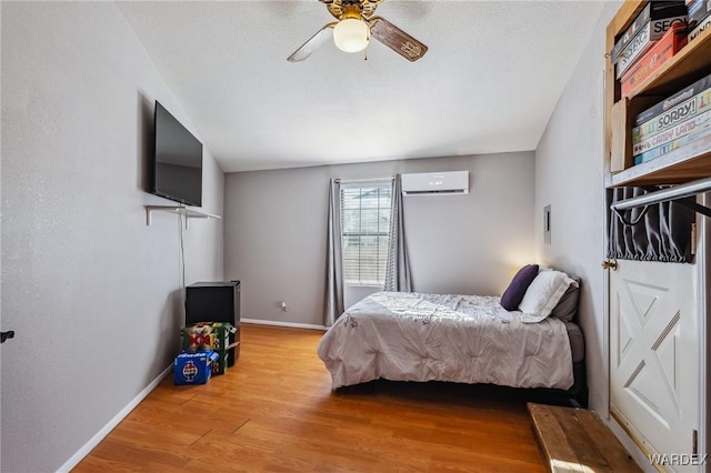 bedroom featuring light wood finished floors, baseboards, ceiling fan, a wall mounted air conditioner, and a textured ceiling