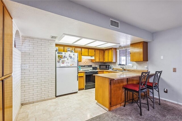 kitchen featuring a peninsula, range with gas stovetop, light countertops, freestanding refrigerator, and brown cabinets