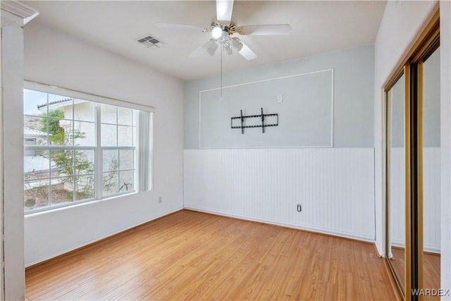 empty room with ceiling fan, light wood-style flooring, a wainscoted wall, and visible vents