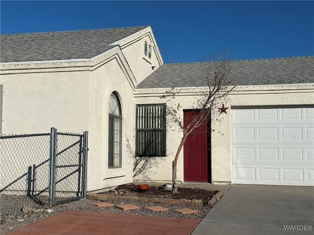 property entrance featuring a shingled roof, fence, concrete driveway, a gate, and stucco siding