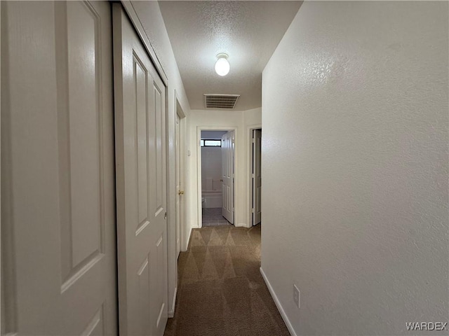 corridor featuring a textured ceiling, a textured wall, visible vents, baseboards, and dark carpet