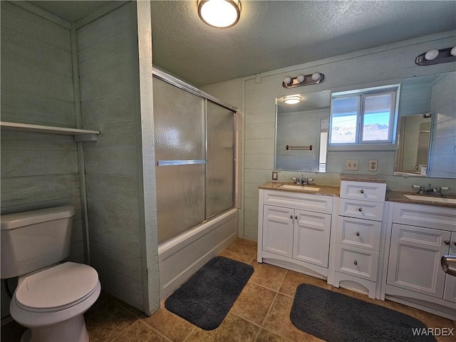 full bath with a textured ceiling, toilet, bath / shower combo with glass door, a sink, and double vanity