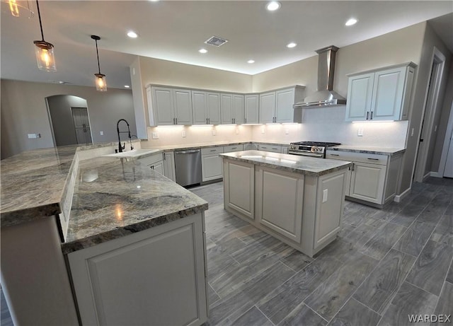 kitchen with arched walkways, stainless steel appliances, a kitchen island, wall chimney range hood, and a peninsula