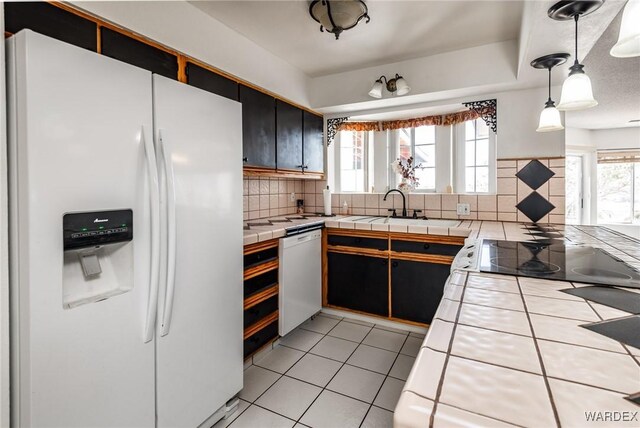 kitchen featuring white refrigerator with ice dispenser, dishwasher, tile countertops, hanging light fixtures, and dark cabinetry