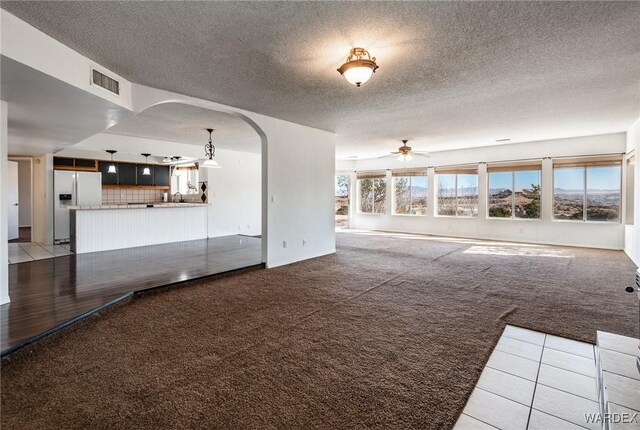 unfurnished living room featuring carpet floors, arched walkways, visible vents, a ceiling fan, and a textured ceiling