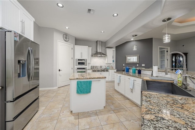 kitchen with decorative light fixtures, appliances with stainless steel finishes, white cabinets, a kitchen island, and wall chimney range hood