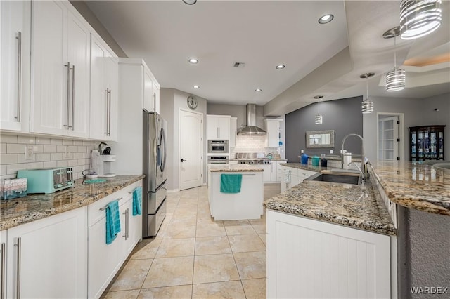 kitchen with decorative light fixtures, a kitchen island with sink, a sink, wall chimney range hood, and white cabinetry