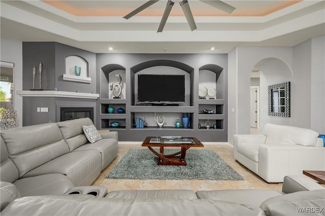 living room with arched walkways, a tray ceiling, built in shelves, and a glass covered fireplace