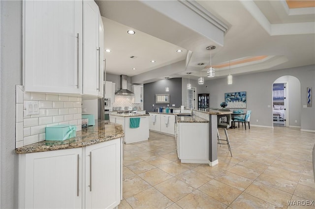 kitchen featuring a center island with sink, a raised ceiling, wall chimney exhaust hood, hanging light fixtures, and white cabinetry