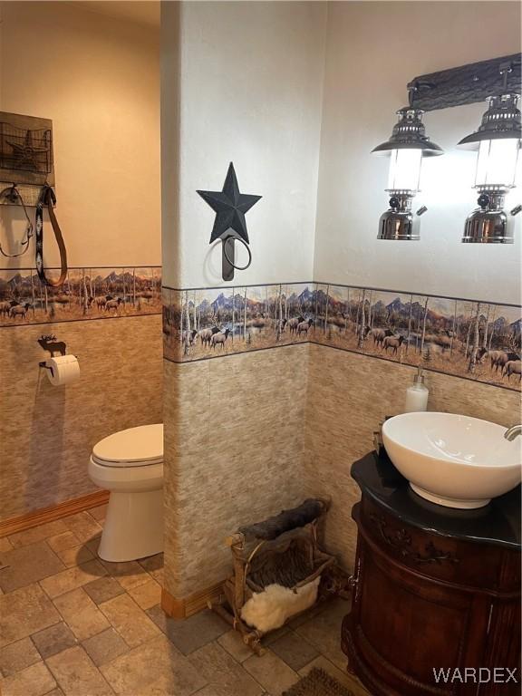 half bath with wainscoting, toilet, stone tile flooring, vanity, and tile walls