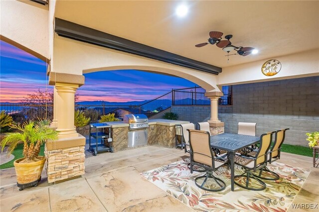 patio terrace at dusk with grilling area, outdoor dining space, ceiling fan, fence, and exterior kitchen