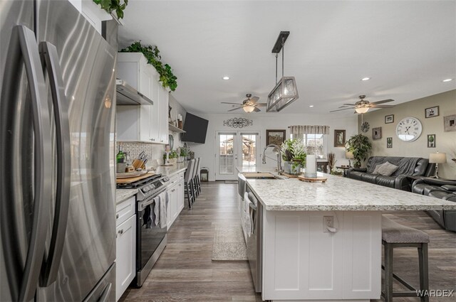 kitchen with a kitchen island with sink, stainless steel appliances, under cabinet range hood, white cabinetry, and pendant lighting