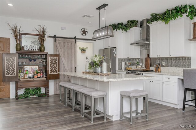 kitchen with a center island with sink, hanging light fixtures, appliances with stainless steel finishes, white cabinets, and wall chimney range hood