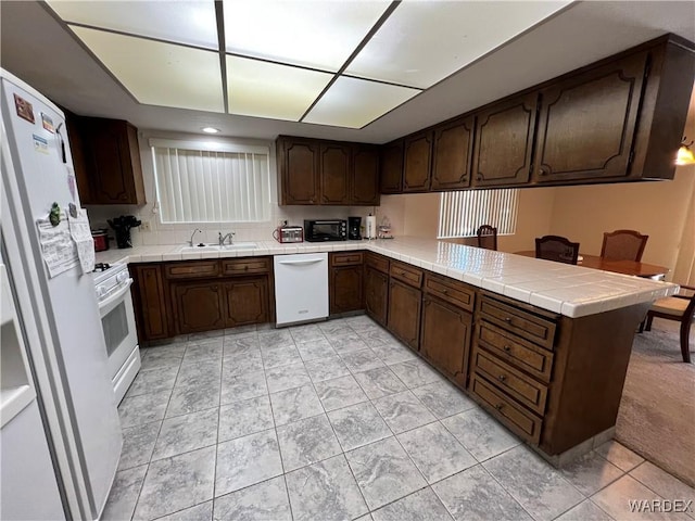 kitchen featuring white appliances, dark brown cabinetry, tile counters, a peninsula, and a sink