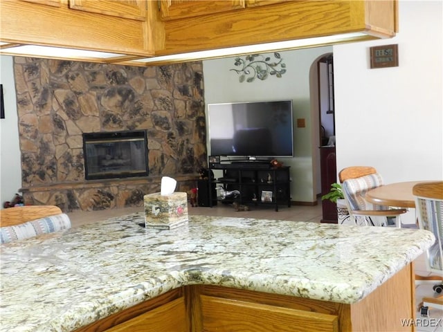 kitchen featuring arched walkways, a peninsula, a fireplace, light stone countertops, and brown cabinetry