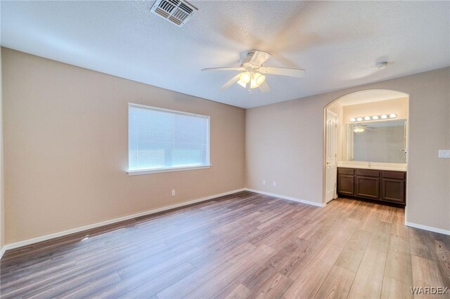 unfurnished bedroom featuring arched walkways, visible vents, baseboards, light wood finished floors, and ensuite bath