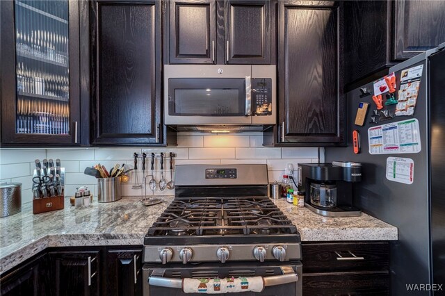 kitchen featuring dark brown cabinets, appliances with stainless steel finishes, light stone countertops, tasteful backsplash, and glass insert cabinets