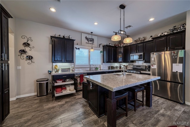 kitchen with an island with sink, appliances with stainless steel finishes, dark wood-type flooring, light stone countertops, and pendant lighting