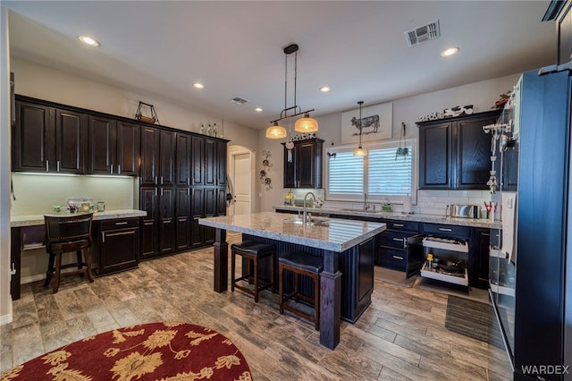 kitchen with light stone counters, a center island with sink, a breakfast bar area, visible vents, and hanging light fixtures