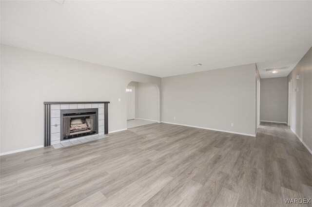 unfurnished living room featuring arched walkways, light wood finished floors, a tiled fireplace, and baseboards