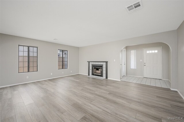 unfurnished living room with arched walkways, a tile fireplace, visible vents, baseboards, and light wood-style floors