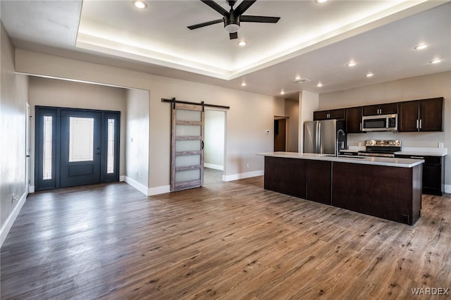 kitchen with a tray ceiling, light countertops, a barn door, appliances with stainless steel finishes, and a kitchen island with sink